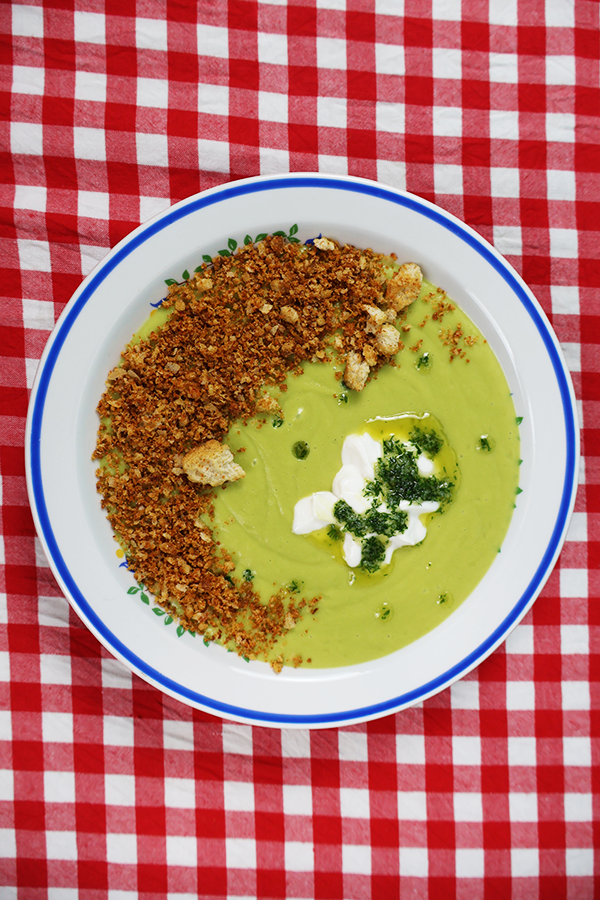 Biało-czerwony obrus w kratkę a na nim biały talerz z niebieską obramówką. W talerzu zielona zupa krem z dodatkiem śmietany, masła koperkowego i prażonego pokruszonego chleba.