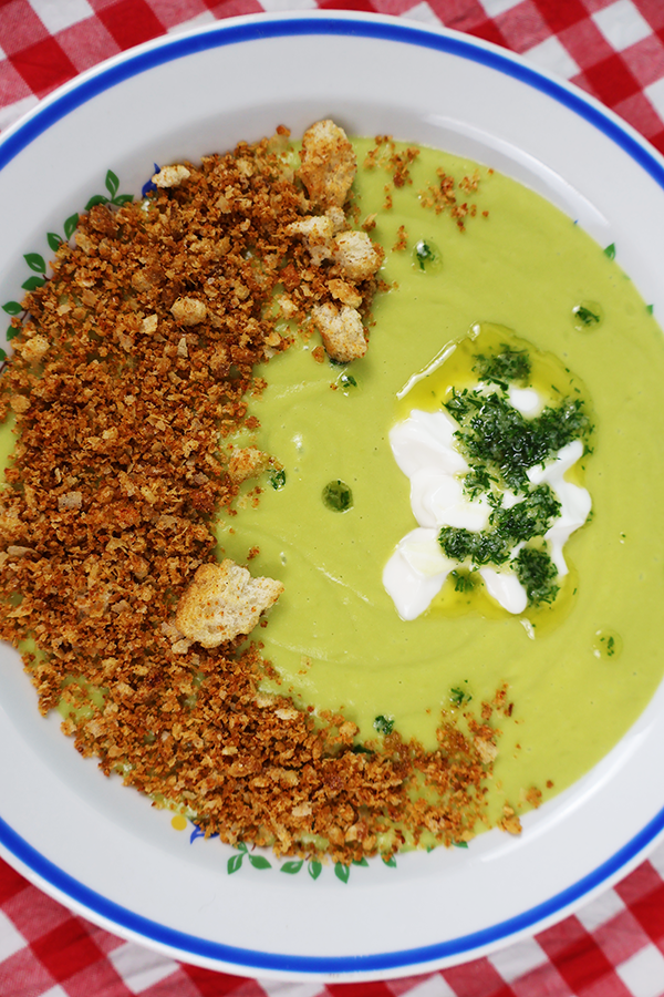 Zbliżenie na biały talerz z niebieską obramówką. W talerzu zielona zupa krem z dodatkiem śmietany, masła koperkowego i prażonego pokruszonego chleba.