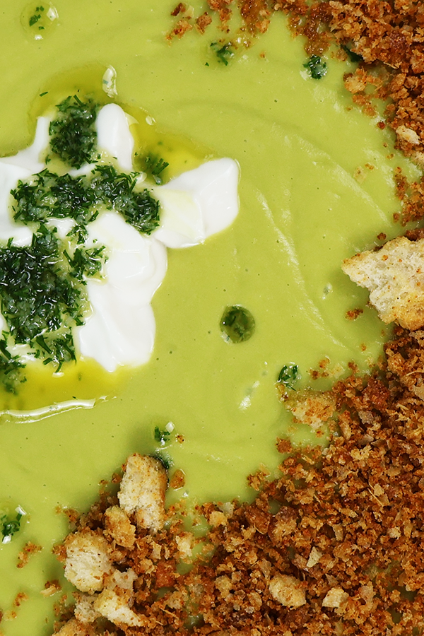 Zbliżenie na zieloną zupę krem z dodatkiem śmietany, masła koperkowego i prażonego pokruszonego chleba.