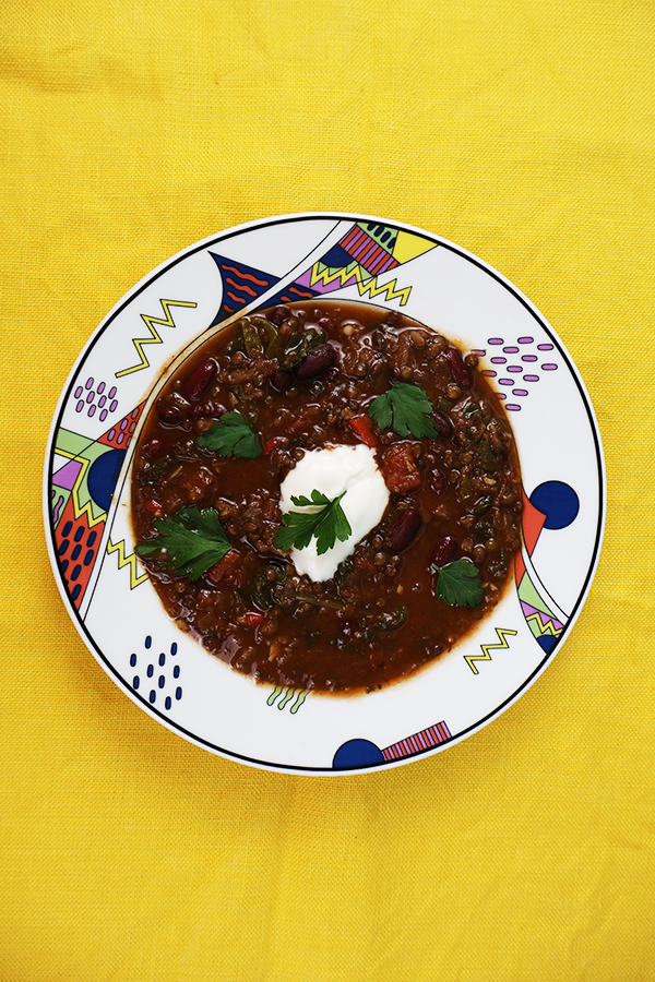 Żółty obrus a na nim kolorowy talerz. W talerzu ciemna zupa z czarną soczewicą, fasolą, śmietaną i pietruszką.
