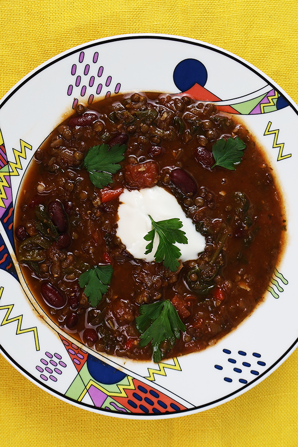 Zbliżenie na kolorowy talerz. W talerzu ciemna zupa z czarną soczewicą, fasolą, śmietaną i pietruszką.