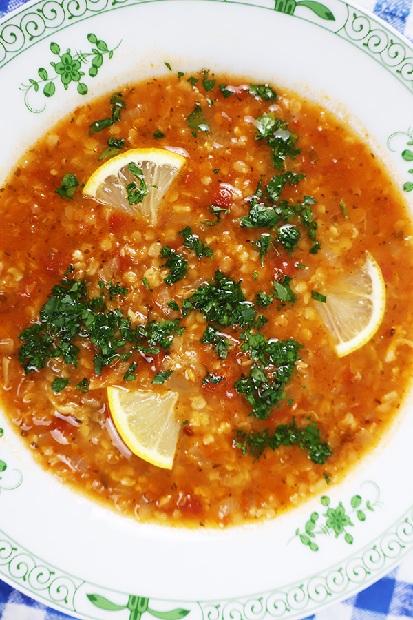 Zbliżenie na biały talerz z zielonymi dodatkami. W talerzu mocno czerwona zupa, z widoczną kaszą bulgur, kawałkami cytryny i posiekaną natką pietruszki.