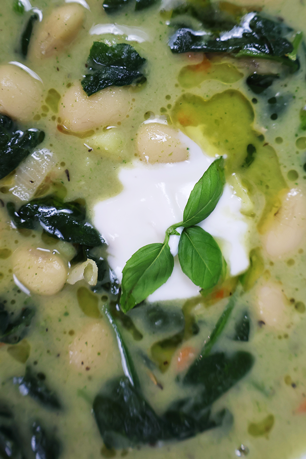 Zbliżenie na zieloną zupę fasolową z dodatkiem szpinaku, śmietany, oleju i listkiem bazylii.