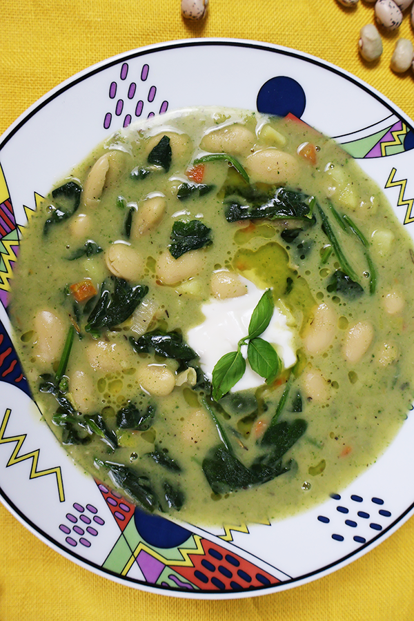 Zbliżenie na kolorowy talerz. W talerzu zielona zupa fasolowa z dodatkiem szpinaku, śmietany, oleju i listkiem bazylii.