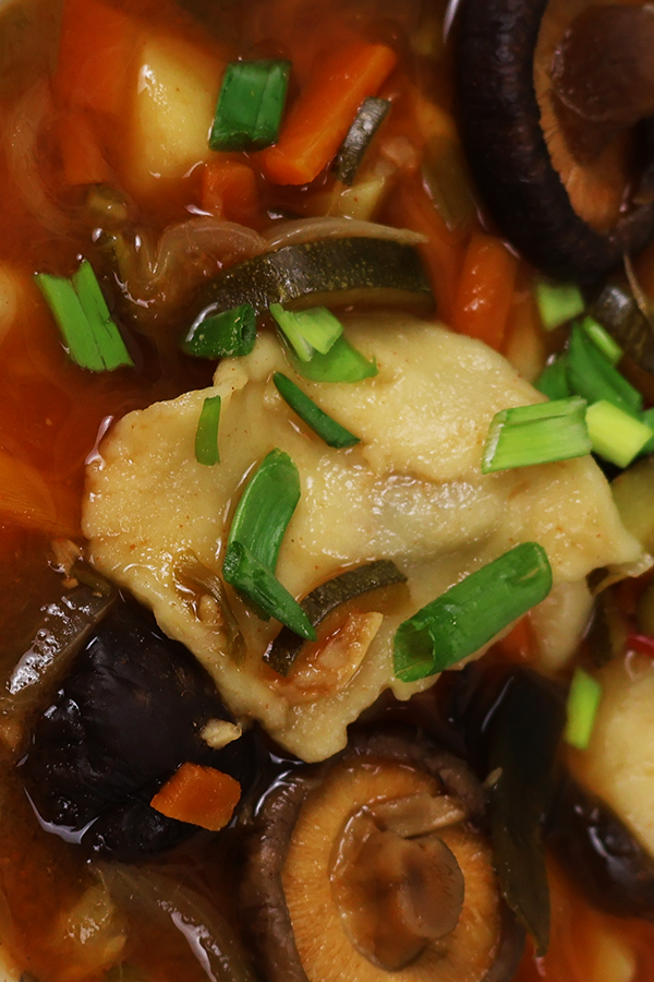 Zbliżenie na ciemną zupę z dodatkiem klusek, cebuli, cukinii, grzybów, marchewki i szczypiorku.