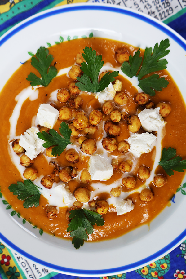 Zbliżenie na biały talerz z niebieską obramówką. W talerzu czerwona zupa z dodatkiem ciecierzycy, śmietany i pietruszki.