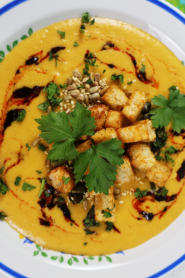 Zbliżenie na biały talerz z niebieską obramówką. W talerzu żółta zupa krem, z dodatkiem grzanek, pestek słonecznika, kolendry i ciemnego sosu.