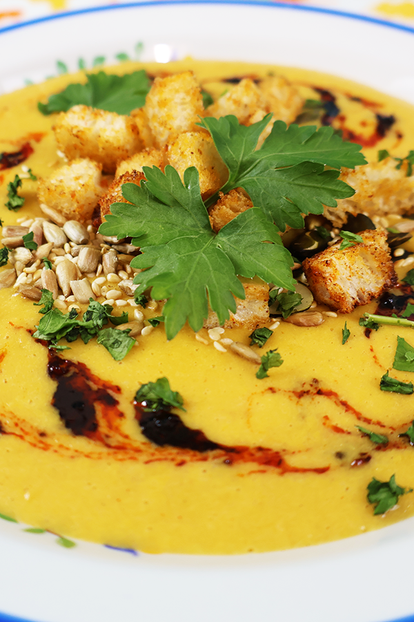 Zbliżenie na biały talerz z niebieską obramówką. W talerzu żółta zupa krem, z dodatkiem grzanek, pestek słonecznika, kolendry i ciemnego sosu.