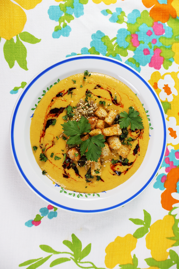 Biały obrus w kolorowe kwiatki, a na nim biały talerz z niebieską obramówką. W talerzu żółta zupa krem, z dodatkiem grzanek, pestek słonecznika, kolendry i ciemnego sosu.