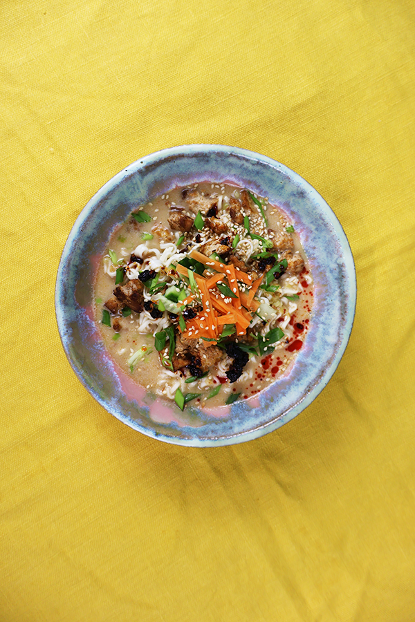 Żółty obrus a na nim szaro-różowy talerz. W talerzu zupa z dodatkiem ogórka, marchewki, tofu, szczypiorku, papryki, sezamu i oleju chili.
