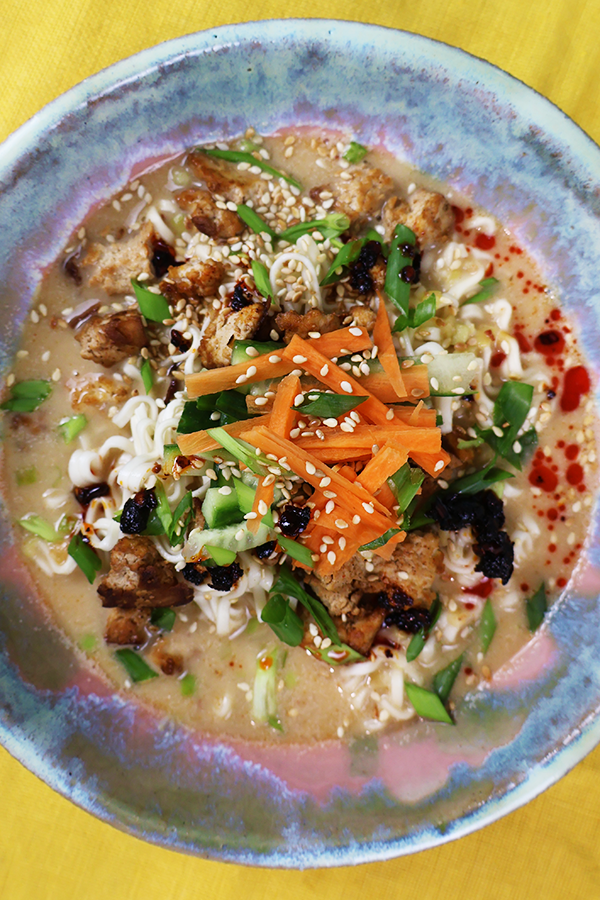 Zbliżenie na szaro-różowy talerz. W talerzu zupa z dodatkiem ogórka, marchewki, tofu, szczypiorku, papryki, sezamu i oleju chili.