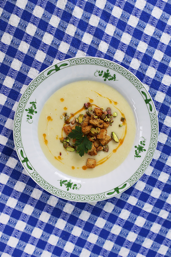 Niebiesko-biały obrus z białym talerzem z zielonymi akcentami. W talerzu kremowa zupa z dodatkiem pikantnej oliwy, grzanek, pistacji i natki pietruszki.