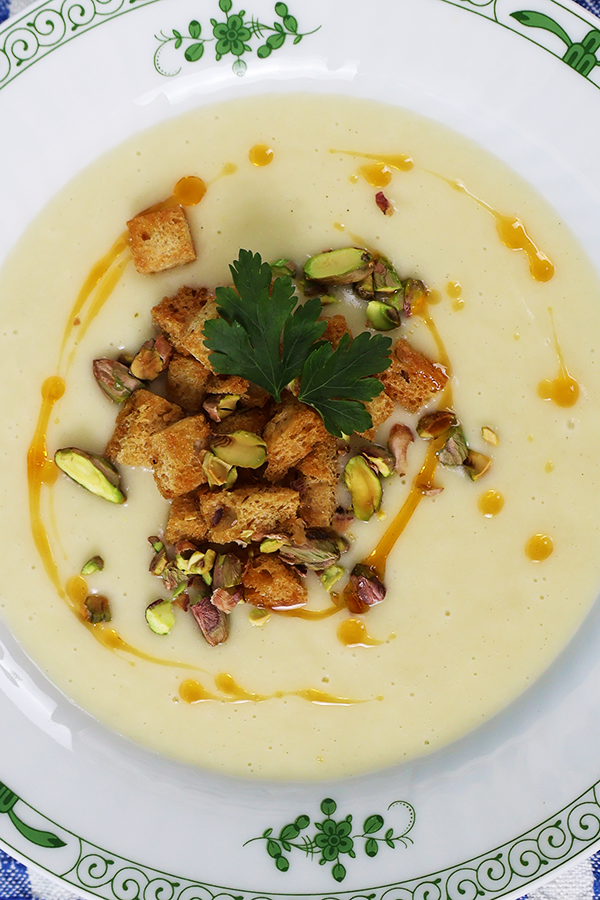 Zbliżenie na talerz z zielonymi akcentami. W talerzu kremowa zupa z dodatkiem pikantnej oliwy, grzanek, pistacji i natki pietruszki.