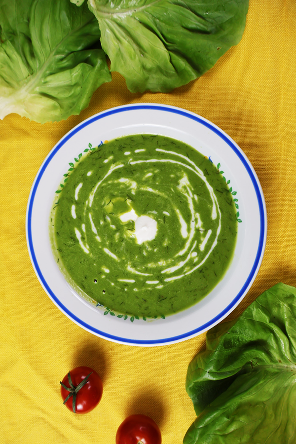 Żółty obrus udekorowany sałatą i pomidorami. Na obrusie biały talerz z niebieską obramówką. W talerzu zielona zupa z dodatkiem śmietany.