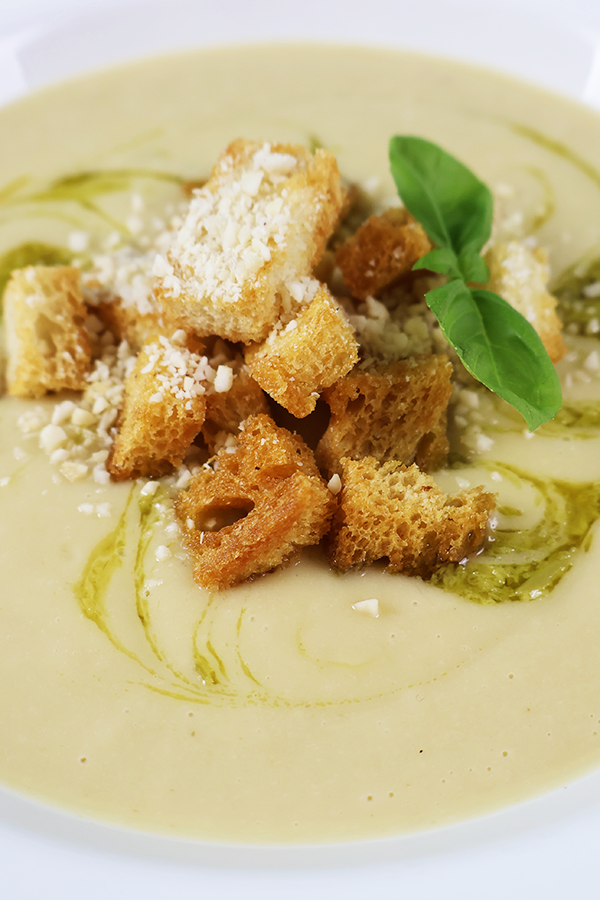 Zbliżenie na kremową zupę z dodatkiem grzanek, oliwy, bazylii i kruszonych migdałów.