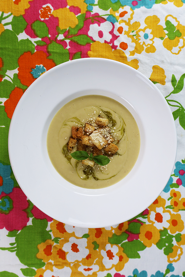 Biały obrus w kolorowe kwiatki a na nim biały talerz. W talerzu kremowa zupa z dodatkiem grzanek, oliwy, bazylii i kruszonych migdałów.