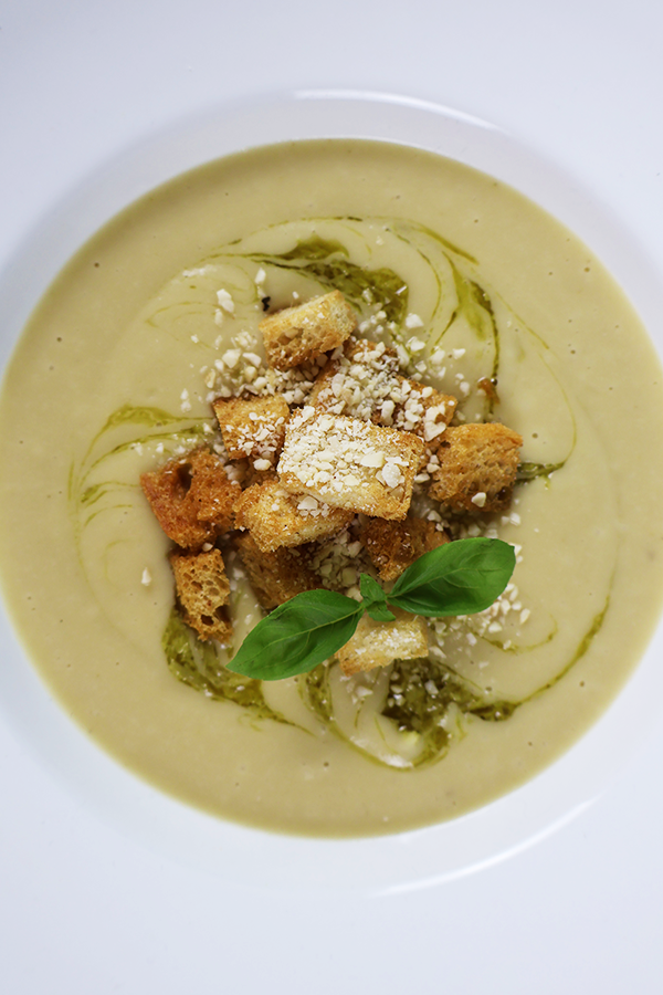 Zbliżenie na biały talerz. W talerzu kremowa zupa z dodatkiem grzanek, oliwy, bazylii i kruszonych migdałów.