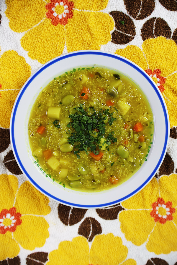 Biały obrus w żółte kwiaty a na nim biały talerz z niebieską obramówką. W talerzu kolorowa zupa z dodatkiem komosy ryżowej, bobu, ziemniaków i posiekanej pietruszki.