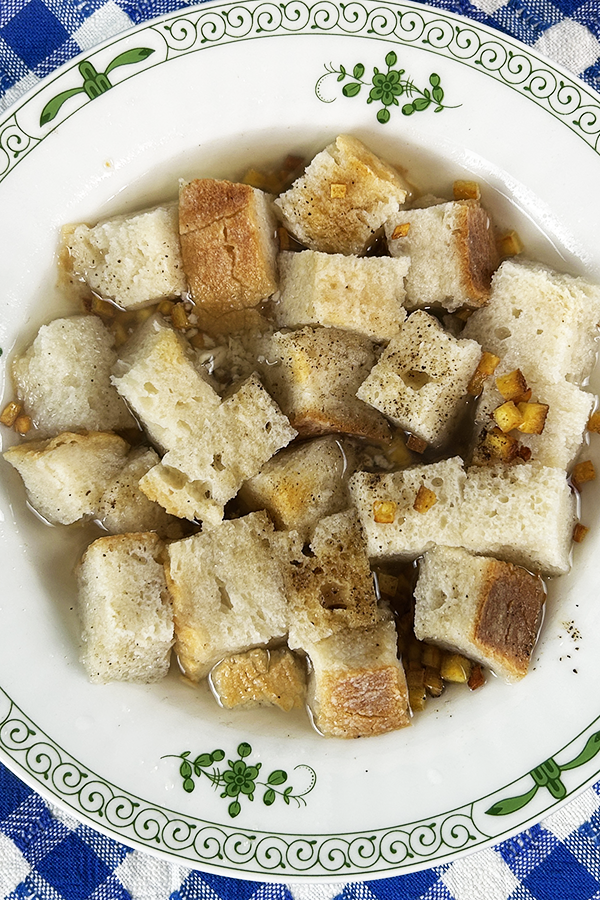 Zbliżenie na biały talerz z zielonymi akcentami. W talerzu kawałki chleba zanurzone w zupie z dodatkiem skwarek z tofu.