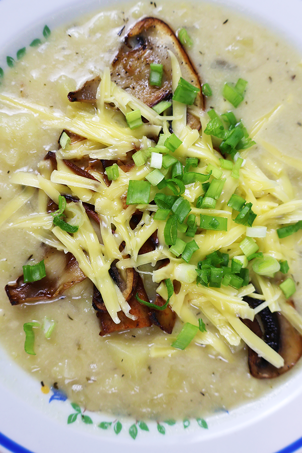 Zbliżenie biały talerz z niebieską obramówką. W talerzu zupa ziemniaczana z dodatkiem pieczarek i sera posypana szczypiorkiem.