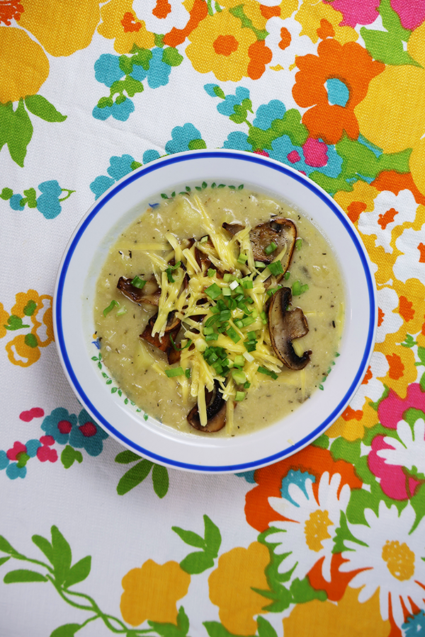 Biały obrus w kolorowe kwiaty a na nim biały talerz z niebieską obramówką. W talerzu zupa ziemniaczana z dodatkiem pieczarek i sera posypana szczypiorkiem.