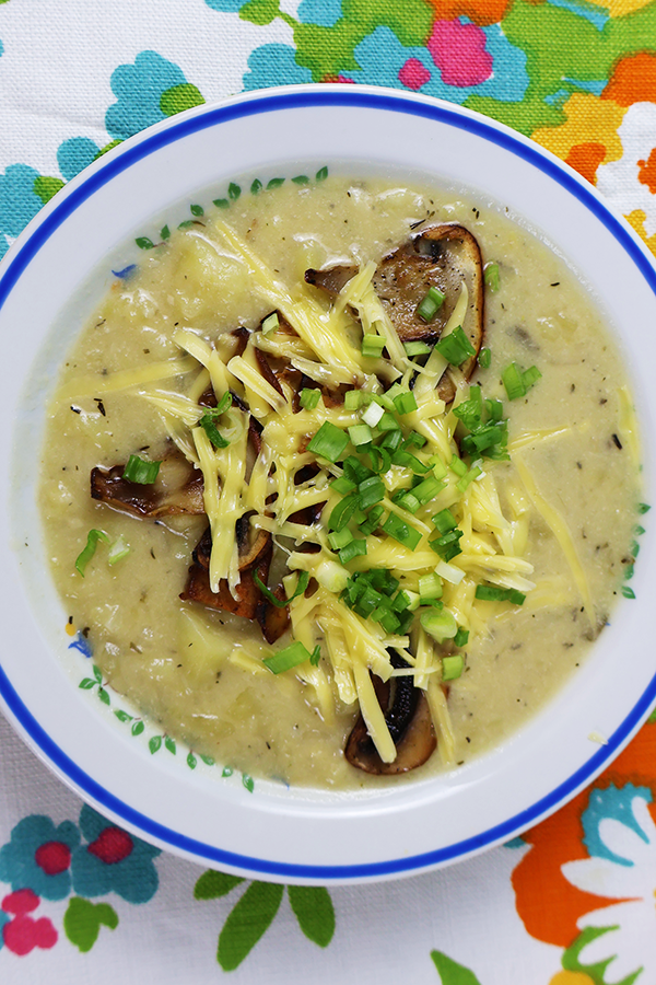 Zbliżenie na biały obrus w kolorowe kwiaty a na nim biały talerz z niebieską obramówką. W talerzu zupa ziemniaczana z dodatkiem pieczarek i sera posypana szczypiorkiem.