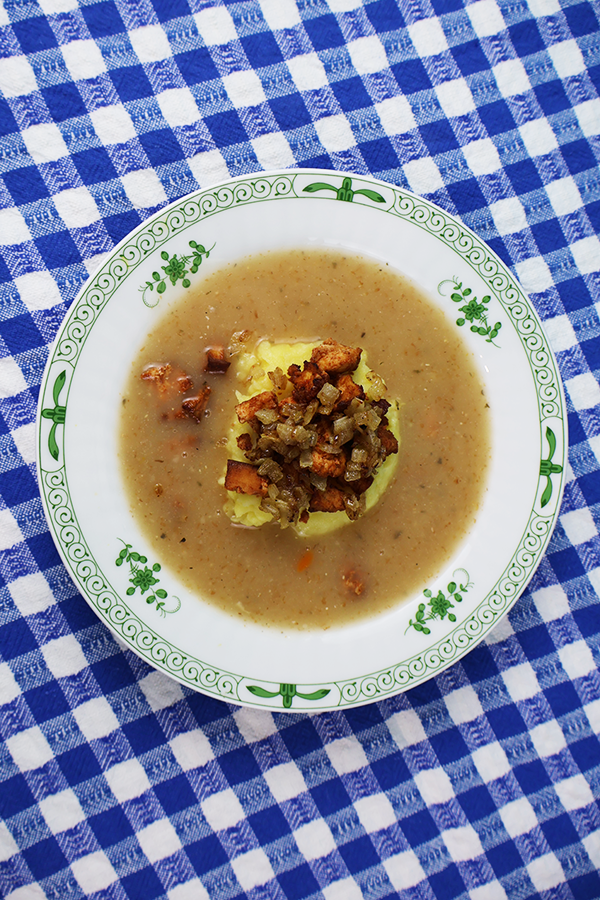 Obrus w biało-niebieską kratkę a na nim biały talerz z zielonymi dodatkami. W talerzu zupa z kopką ziemniaków i skwarkami z tofu i cebuli.