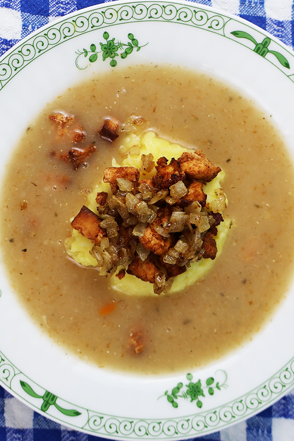 Zbliżenie na biały talerz z zielonymi dodatkami. W talerzu zupa z kopką ziemniaków i skwarkami z tofu i cebuli.